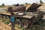 A child standing next to a tank which has been left abandoned and rusting. 